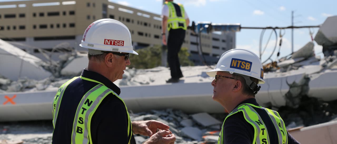 Two NTSB investigators in hard hats and safety vests talk in front of the broken concrete of the collapsed bridge