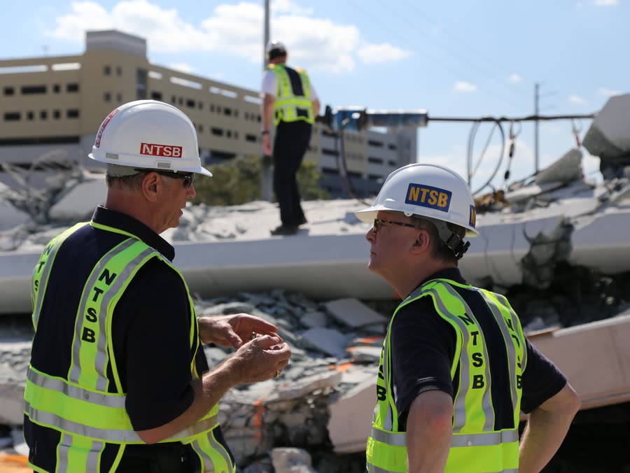 Two NTSB investigators in hard hats and safety vests talk in front of the broken concrete of the collapsed bridge