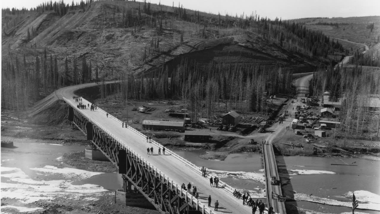 A long timber bridge across the Sikanni Chief River, with a crowd of people walking over it and forested hills behind, 1943